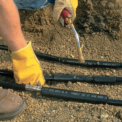 A worker wearing protective gloves is using a heat gun to apply heat shrink tubing around black electrical cables.